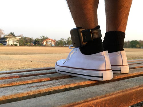 Person seated on an outdoor bench wearing a GPS ankle monitor above a white sneaker