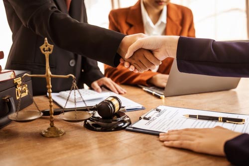 Two people shaking hands across a desk of legal documents, with a scale and gavel nearby